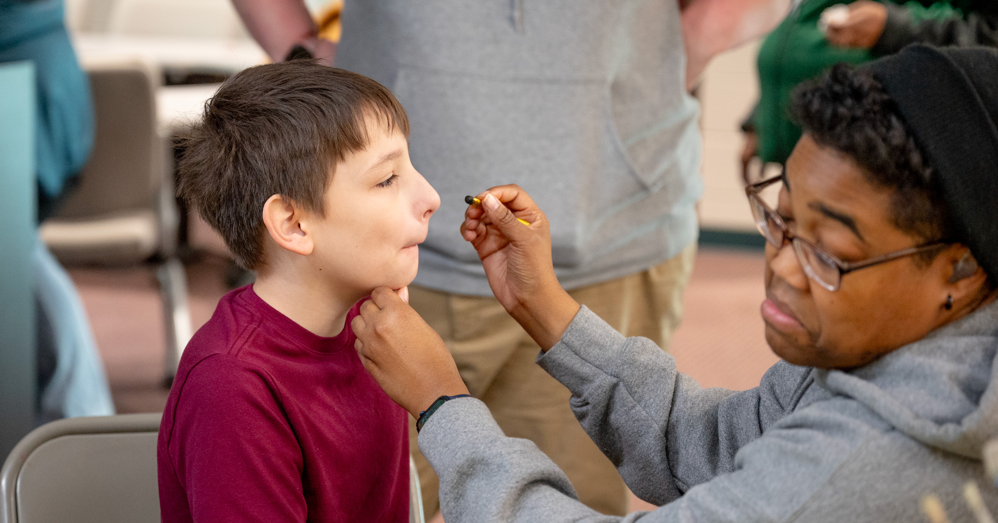 Fall Fest:  Regana holds a face paint crayon and looks at reference as she starts to pain a student's face
